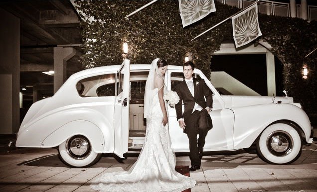 A bride and groom standing in front of an old car.