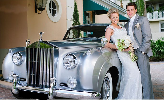 A bride and groom pose in front of a rolls royce.